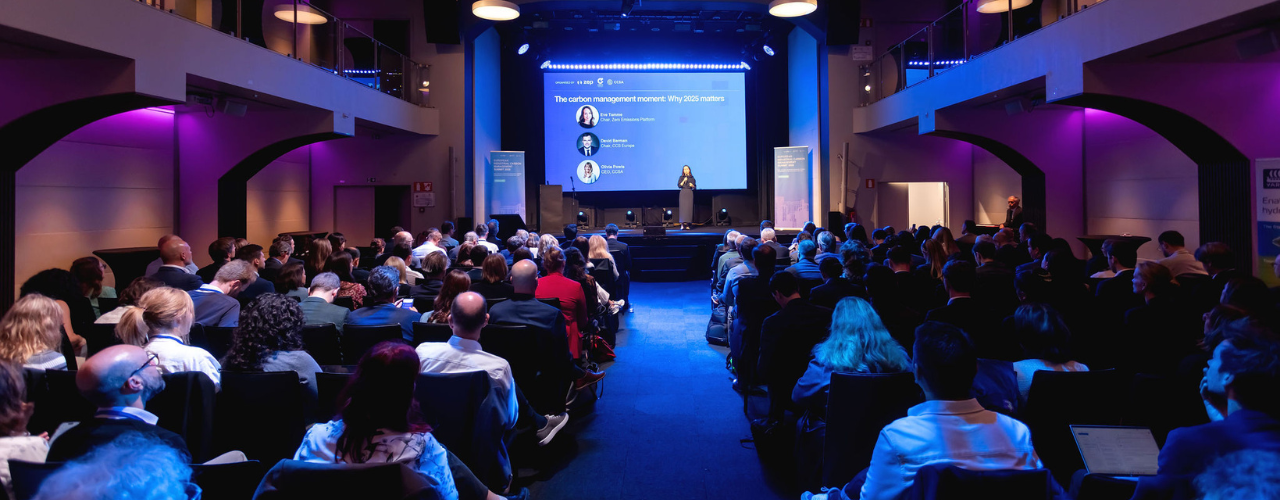 A wide-angle view of a large conference auditorium filled with attendees seated and facing a stage. A speaker stands center stage beneath blue lighting, presenting in front of a large screen displaying a panel session on carbon management and why 2025 matters. Balconies line both sides of the hall, and the audience is attentively listening.