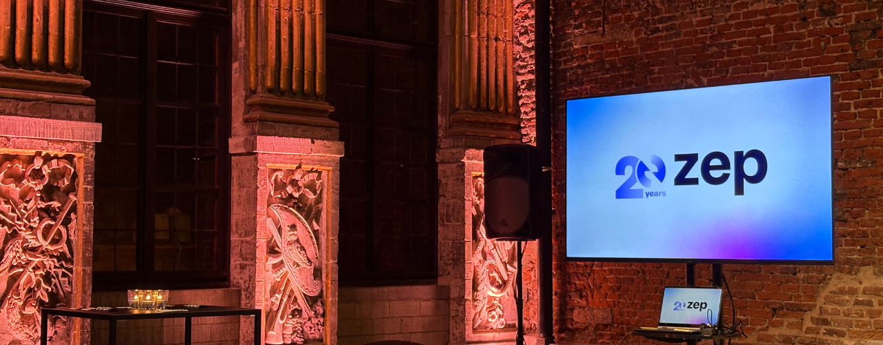 An indoor event space with exposed brick walls and ornate stone columns lit by warm pink and orange lighting. On the right side, a large screen displays the logo “20 years ZEP” against a blue gradient background, with a laptop showing the same logo on a small stand below it. A tall speaker stands between the screen and the columns. On the left, a high table with several lit candles sits in front of the illuminated architectural details.